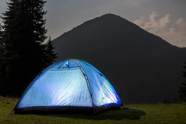 Tourist hikers tent brightly lit from inside on green grassy valley among pine trees under dark blue cloudy sky on distant mountain covered with forest background. Summer night camping in mountains.