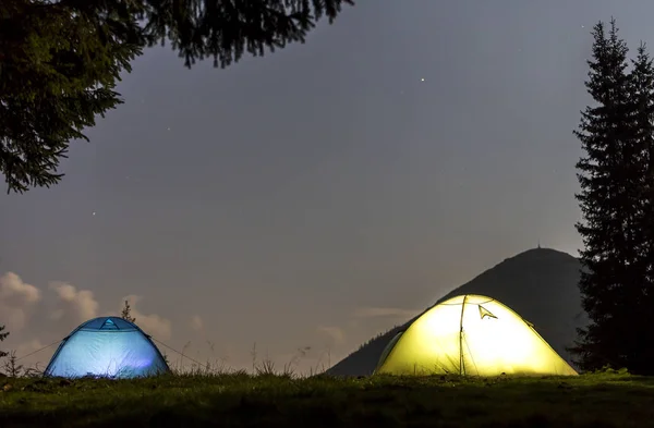 Two brightly lit tourist tents on green grassy forest clearing on dark mountain and clear blue starry sky copy space background. Tourism, night camping in summer mountains, beauty of nature concept.