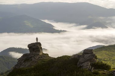 Atletik dağcı turist dağ Vadisi üzerinde yüksek kayalık oluşumu üzerinde silueti beyaz bulutlar ve sis ile dolu ve herdem yeşil orman dağ yamaçları altında açık gökyüzü arka plan ile kaplı.