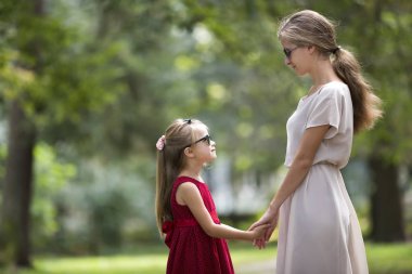Profil genç sarışın uzun saçlı çekici gülümseyen kadın ve kız küçük çocuk güneş gözlüğü ve birbirlerine güneşli yaz yeşil bokeh özgeçmişlerine bakarak elele moda elbiseler.