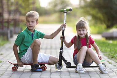 İki sevimli mutlu komik gülümseyen genç çocuk ve kamera için poz scooter ve güneşli renkli arka plan bulanık üzerinde kaykay üzerinde oturan güzel uzun saçlı kız yakışıklı çocuk kız.