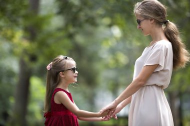 Profil genç sarışın uzun saçlı çekici gülümseyen kadın ve kız küçük çocuk güneş gözlüğü ve birbirlerine güneşli yaz yeşil bokeh özgeçmişlerine bakarak elele moda elbiseler.