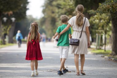 Genç sarışın uzun saçlı kadın iki çocuk, küçük kızı ve oğluyla güneşli park sokak boyunca sıcak yaz gününde bulanık insanlar siluet ve yeşil ağaçlar bokeh arka plan üzerinde yürüme arkadan görünüm.