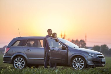 Genç çift, çekici ince uzun - saçlı kadın ve yakışıklı adam birlikte gündoğumu veya günbatımı parlak gökyüzü üzerinde kırsal kırsalında açık kapı ile gümüş renkli araba, ayakta kopyalama alanı arka plan.