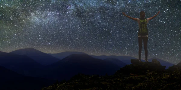 Back view of young slim backpacker tourist girl with raised arms on rocky mountain top on dark night starry sky and foggy mountain range panorama background. Tourism, traveling and climbing concept.