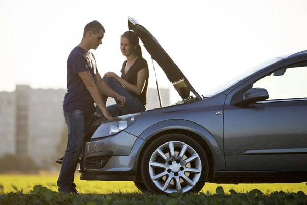 Handsome man at car with popped hood checking oil level in engine using dipstick and attractive woman watching on clear sky background. Transportation, vehicles problems and breakdowns concept.