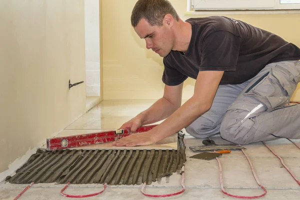 若年労働者の瓦職人が赤い電気ケーブル ワイヤー暖房とセメントの床にレバーを使用してセラミック タイルを取付けます 快適な暖かいホーム コンセプト — ストック写真
