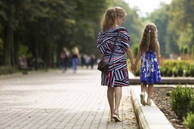 Genç sarışın uzun saçlı ince çekici kadın ve moda elbiseler tutuşup güneşli yaz park uğrayacağız yeşil ağaçlar ve insanlar arka plan bulanık üzerinde yürüyen küçük çocuk kız görünümünü geri.