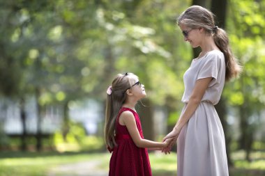 Profil genç sarışın uzun saçlı çekici gülümseyen kadın ve kız küçük çocuk güneş gözlüğü ve birbirlerine güneşli yaz yeşil bokeh özgeçmişlerine bakarak elele moda elbiseler.