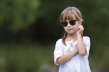 Sevimli çok komik moda moody tatminsiz üzgün kızgın genç kız sarışın örgüler beyaz elbise ve bulanık güneşli yaz yeşil açık havada kopya alan arka plan üzerinde koyu güneş gözlüğü ile portresi.