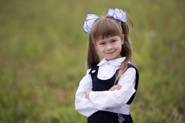 Yakın çekim portre sevimli tatlı gülümseyen emin ilk sınıf öğrencisi kızın okul üniforması ve beyaz yay bulanık açık yeşil güneşli uzun sarı saç alan arka plan açık havada kopyalama.