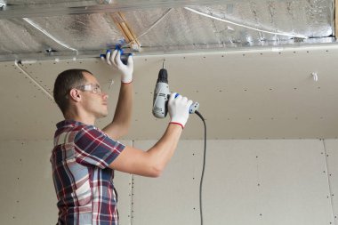 Alçıpan sabitleme Gözlük Genç adam metal çerçeve parlak alüminyum folyo ile yalıtımlı tavanda Elektrikli tornavida kullanarak tavana askıya. Tadilat, inşaat, do It yourself konsepti.