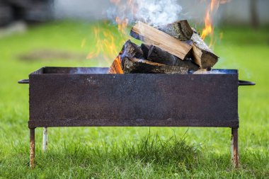 Yakın çekim parlak metal kutuda yanan odun Barbekü güneşli yaz gününde açık için günlüğe kaydeder. Turuncu yüksek alev üzerinde bulanık mavi gökyüzü ve yeşil çim arka plan. Kamp, Emanet ve Turizm kavramı.