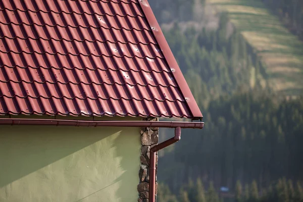 Detail of new modern house cottage corner with stucco walls decorated with natural stones, red shingled roof and rain gutter pipe system on blurred spruce forest copy space background.