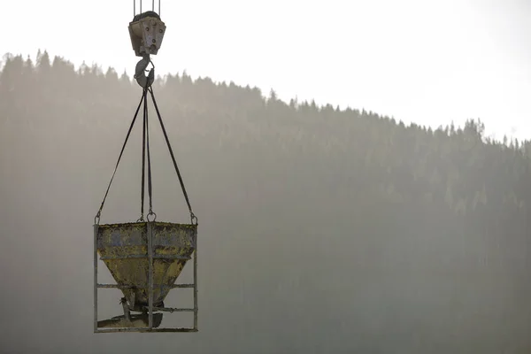 Bucket container with liquid concrete hanging on crane hook on copy space background. Construction machinery equipment.