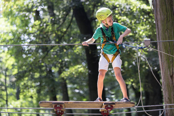 Young cute child boy in summer clothing, safety harness and helmet attached with carbine to cable moves slowly along rope way on green trees sunny bokeh background. Sport, game, leisure concept.