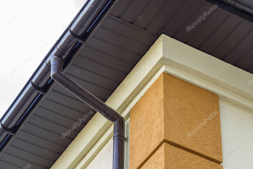 Close-up detail of cottage house corner with brown metal planks siding and roof with steel gutter rain system. Roofing, construction, drainage pipes installation and connection concept.