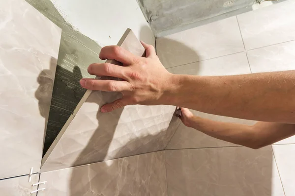 Close-up of worker tiler hands installing light beige ceramic tiles on walls of future bathroom. Tiles installation, home improvement, renovation and construction, comfortable modern home concept.