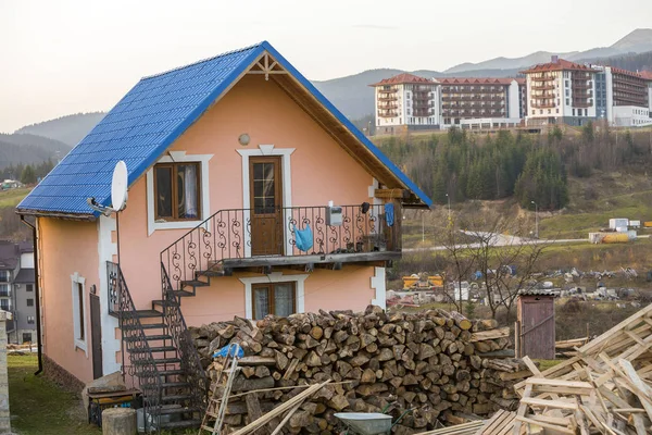 Two-storied residential cottage house with shingle roof, plastered pink walls, forged balcony and outside staircase, pile of firewood in ecological mountain area on sunny summer day.