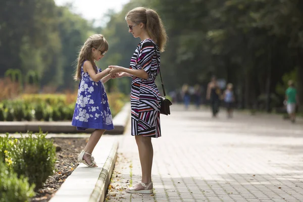 Genç sarışın uzun saçlı çekici kadın ve kız küçük çocuk güneş gözlüğü ve şık elbiseler güneşli park sokak üzerinde el ele tutuşarak profili tam uzunlukta portresi. Mutlu aile ilişkileri kavramı.