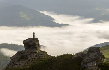 Atletik dağcı turist dağ Vadisi üzerinde yüksek kayalık oluşumu üzerinde silueti beyaz bulutlar ve sis ile dolu ve herdem yeşil orman dağ yamaçları altında açık gökyüzü arka plan ile kaplı.