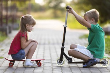 İki sevimli mutlu komik gülümseyen genç çocuk ve kamera için poz scooter ve güneşli renkli arka plan bulanık üzerinde kaykay üzerinde oturan güzel uzun saçlı kız yakışıklı çocuk kız.