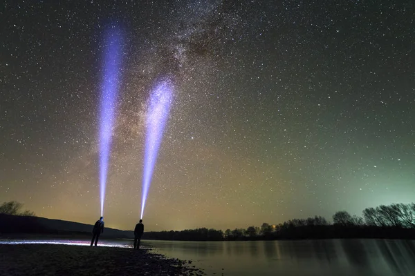 Back view of two men with head flashlight standing on river bank, long blue beam across beautiful dark starry sky. Night photography concept. Wide panorama, copy space background.