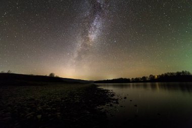 Çakıl taşları nehir banka, ağaçlar ufuk, parlak yıldızlar ve Samanyolu karanlık gökyüzü manzara sessiz suya yansıyan. Doğa kavramının güzellik.