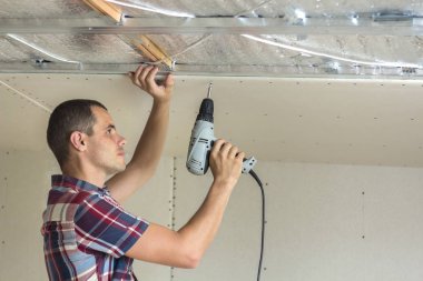 Her zamanki giyim ve iş eldivenleri Alçıpan sabitleme genç adam metal çerçeve parlak alüminyum folyo ile yalıtımlı tavanda Elektrikli tornavida kullanarak tavana askıya. DIY, kendiniz yapın kavramı.