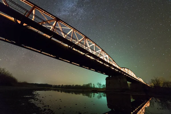 Illuminated metal bridge on concrete supports reflected in water on dark starry sky with Milky Way constellation background. Night photography concept.