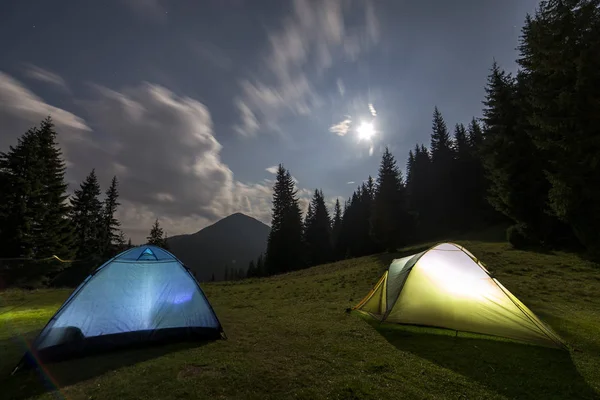 Bright big moon in dark blue cloudy sky over two tourist tents on green grassy forest clearing among tall pine trees on distant mountain background. Tourism, night camping in summer mountains.