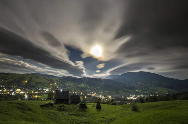 Summer night mountain panorama. Old wooden weathered shepherd huts on green clearing on dramatic cloudy evening sky background, bright road with moving cars and dwelling lights below in valley.