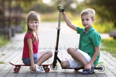 İki sevimli mutlu komik gülümseyen genç çocuk ve kamera için poz scooter ve güneşli renkli arka plan bulanık üzerinde kaykay üzerinde oturan güzel uzun saçlı kız yakışıklı çocuk kız.