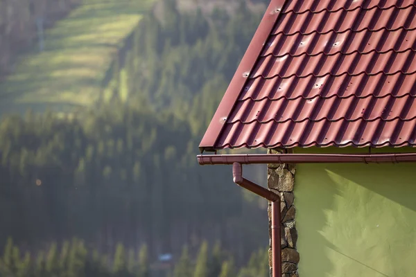 Detail of new modern house cottage corner with stucco walls decorated with natural stones, red shingled roof and rain gutter pipe system on blurred spruce forest copy space background.