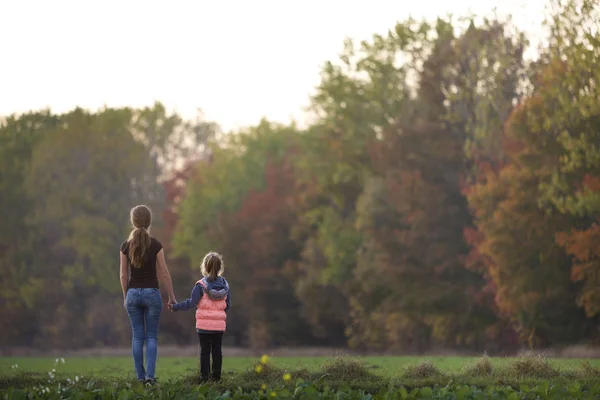 Baksidan på ung smal attraktiva mor och barn flicka stående i grön äng hålla händer utomhus på skogens träd suddig bakgrund. — Stockfoto