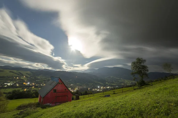 Noche de verano panorama de montaña. Pequeña casa de campo en pendiente de montaña empinada en el fondo dramático cielo de la noche, carretera brillante con coches en movimiento y luces de vivienda del complejo en el valle . — Foto de Stock