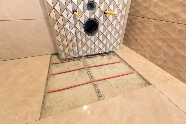 Unfinished reconstruction of bathroom with ceramic tiles installed on walls, heating electrical cables system on cement floor and place for toilet.