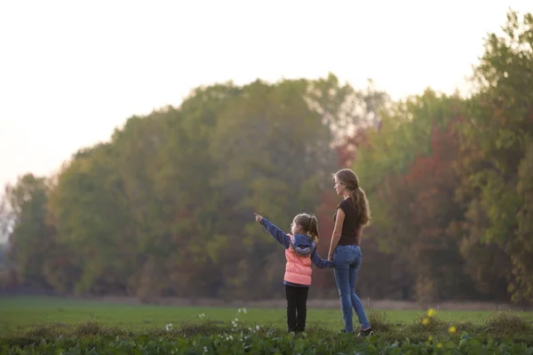 Niña pequeña apunta a distancia cogida de la mano de la atractiva madre en el prado verde al aire libre disfrutando de la naturaleza en los árboles del bosque fondo borroso. Concepto de relaciones familiares felices . — Foto de Stock