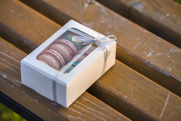 Caja de regalo blanca de cartón con coloridas galletas macaron hechas a mano en un banco de madera al aire libre . — Foto de Stock