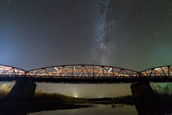 Ponte de metal iluminada em suportes de concreto refletida na água no céu estrelado escuro com fundo constelação Via Láctea. Conceito de fotografia noturna . — Fotografia de Stock