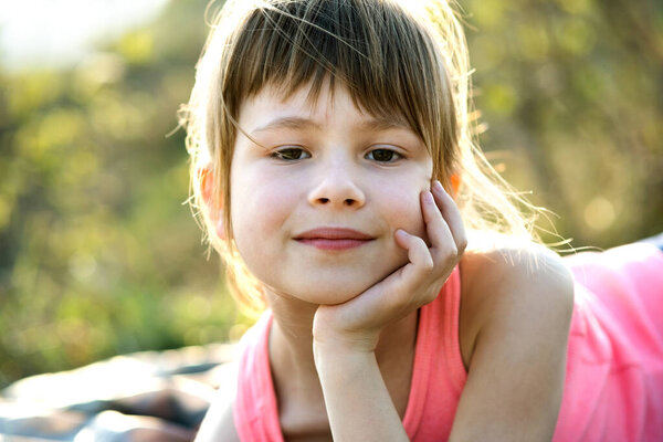 Portrait of pretty child girl with gray eyes and long fair hair leaning on her hands smiling happily outdoors on blurred bright background. Cute female kid on warm summer day outside.