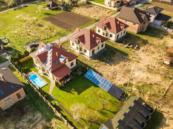Aerial view of a private house with green grass covered yard, solar panels on roof, swimming pool with blue water and wind turbine generator.