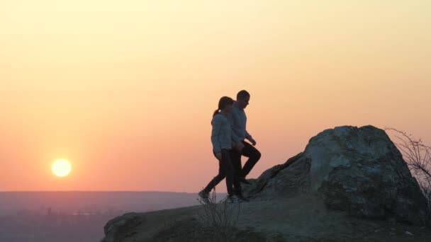 男と女のハイカーは山の中で日没時に石を登るのを助け合っています 夫婦は夜の自然界で高い岩の上に登る 健康的なライフスタイルの概念 — ストック動画