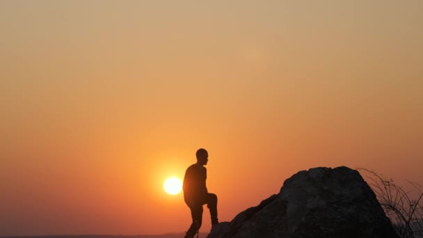 山の中で日没の大きな石の上に一人で登山男ハイカーのシルエット 夕方の自然界の高い岩の上に座っている女性観光客 健康的なライフスタイルの概念 — ストック動画
