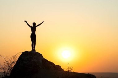 Gün batımında dağlarda büyük bir taşın üzerinde tek başına duran bir kadın yürüyüşçünün silueti. Kadın turist, akşam vakti elini yüksek kayalara kaldırıyor. Turizm, seyahat ve sağlıklı yaşam tarzı konsepti.