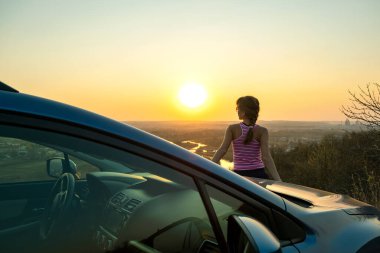 Arabasının yanında duran genç kadın sürücü sıcak günbatımı manzarasının keyfini çıkarıyor. Kız gezgin gece ufkuna bakarak araç kaputuna yaslanıyor..