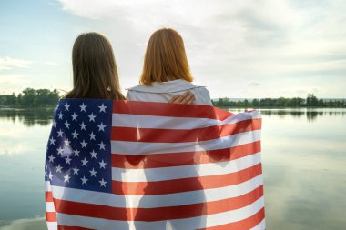 Omuzlarında ABD bayrağı olan iki genç arkadaş göl kıyısında kucaklaşıyorlar. Vatansever kızlar Amerika 'nın bağımsızlık gününü kutluyor.