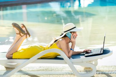 Plaj koltuğunda uzanmış, bilgisayarla çalışan, kablosuz internete bağlı, yaz tatilinde cep telefonuyla konuşan genç bir kadın. Seyahat sırasında iş yapmak.