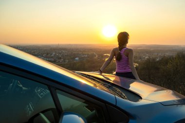 Arabasının yanında duran genç kadın sürücü sıcak günbatımı manzarasının keyfini çıkarıyor. Kız gezgin gece ufkuna bakarak araç kaputuna yaslanıyor..