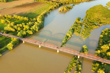 Yeşil kırsal bölgedeki çamurlu geniş nehir üzerinde uzanan dar bir yol köprüsünün havadan görünüşü.